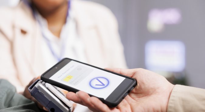 Close up of male customer holding smartphone making nfc contactless payment with electronic credit card, paying for clothes at cashier desk, selective focus. Shopper using mobile payment app